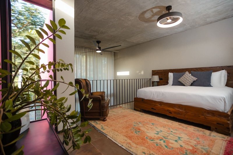 Loft style bedroom with a wooden bed white linens gray pillows a brown leather chair potted plant and a colorful rug