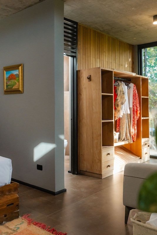 Open wooden wardrobe with hanging clothes and built in shelves in a sunlit room next to a gray wall with a small framed painting
