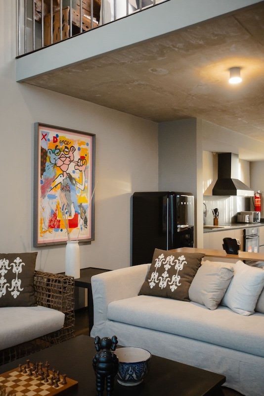 Open plan living area with a grey sofa patterned cushions a chessboard on a dark coffee table and a colorful abstract painting on the wall