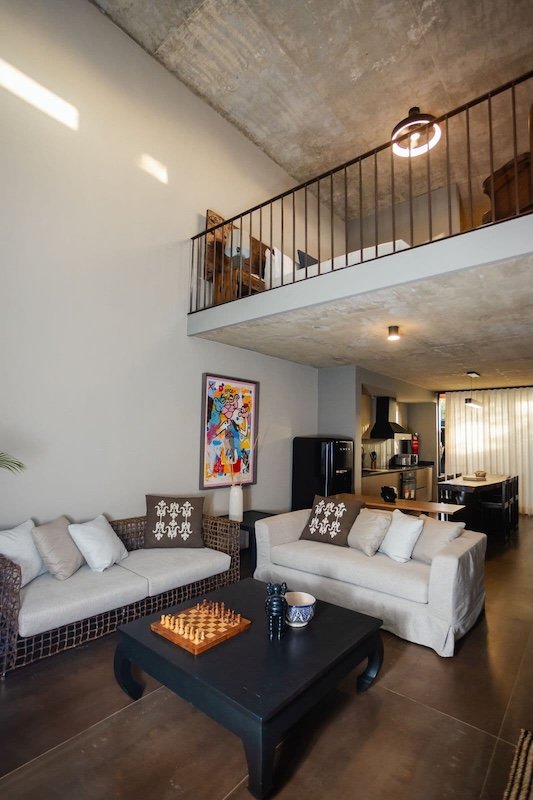 Open concept loft living area with beige sofas a black coffee table featuring a chessboard and colorful wall art mezzanine balcony above