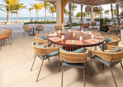 Outdoor beachside restaurant with a round wooden table and wicker chairs, palm trees and the ocean in the background.