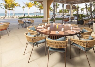 Outdoor beachside restaurant with a round wooden table and wicker chairs, palm trees and the ocean in the background.
