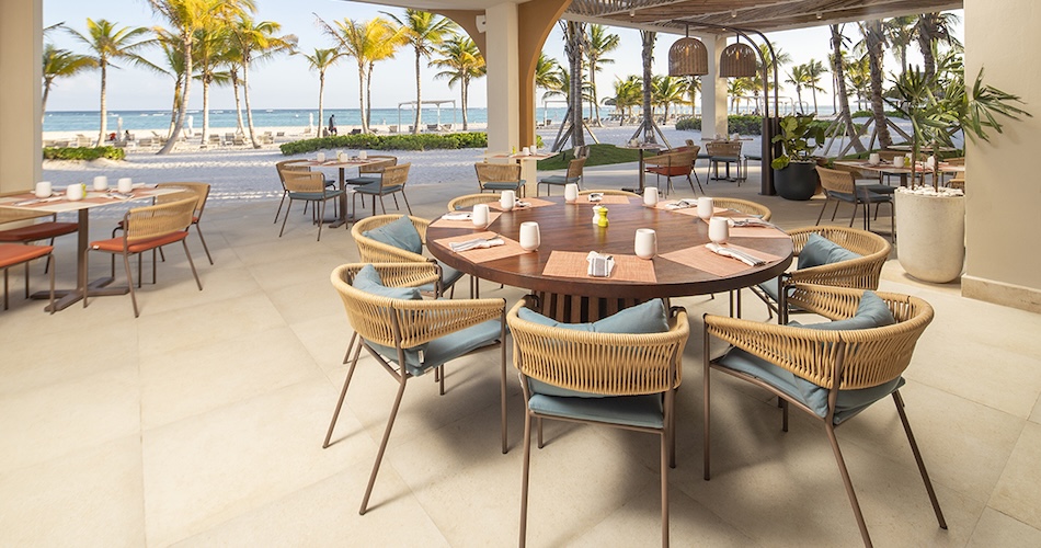 Outdoor beachside restaurant with a round wooden table and wicker chairs palm trees and the ocean in the background