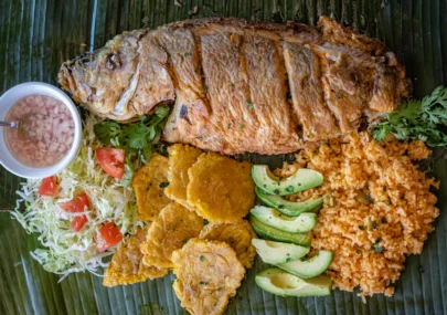 Grilled fish with sides: rice, avocado slices, fried plantains, shredded cabbage with tomato, cilantro, and a small bowl of relish on a banana leaf surface.