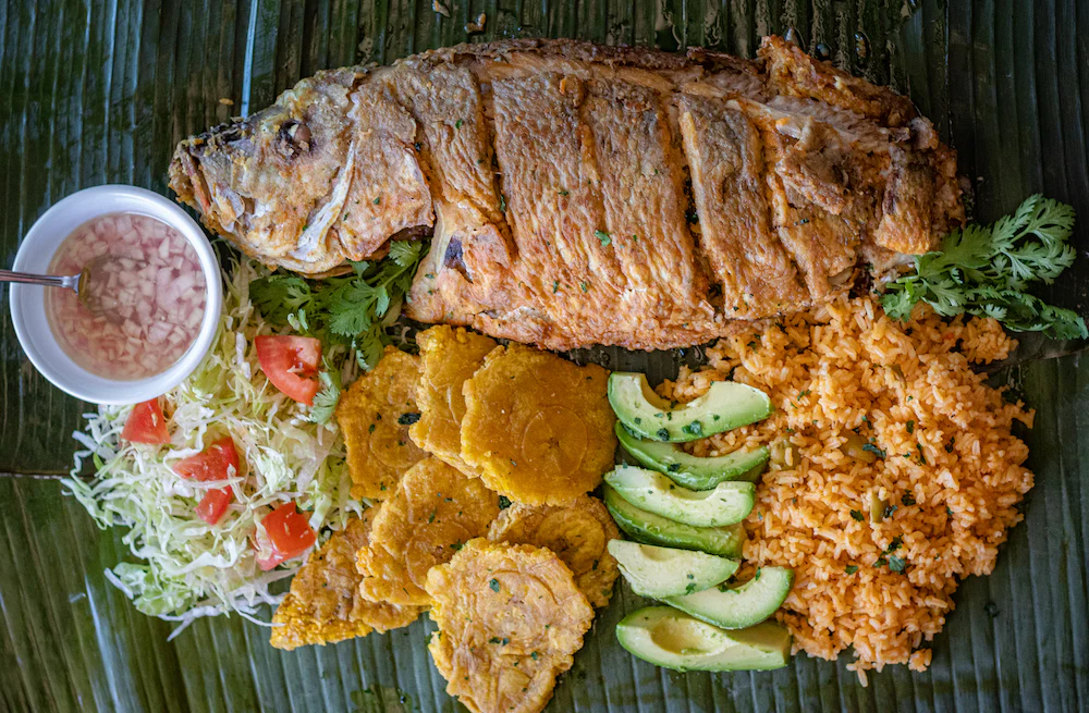 Grilled fish with sides rice avocado slices fried plantains shredded cabbage with tomato cilantro and a small bowl of relish on a banana leaf surface