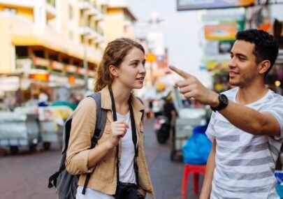 Two young adults chat on a busy street market; man points as the woman listens, camera slung around her neck and backpack on her shoulders.