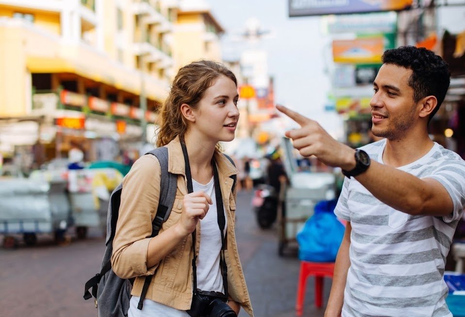 Two young adults chat on a busy street market man points as the woman listens camera slung around her neck and backpack on her shoulders