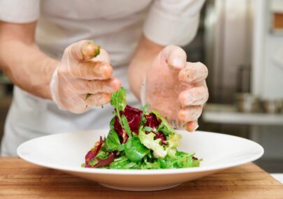 Gloved hands arranging a mixed green salad on a white plate in a kitchen.
