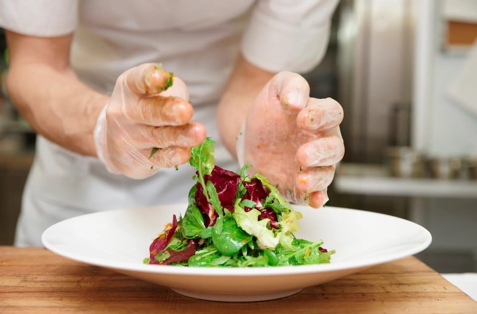 Gloved hands arranging a mixed green salad on a white plate in a kitchen