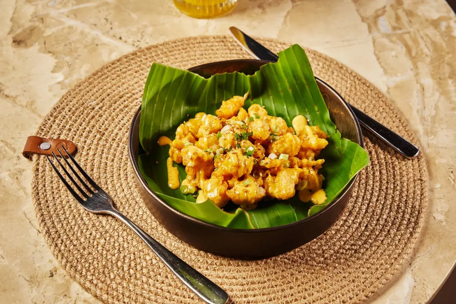 Cabanna the hidden inn punta cana crispy fried chicken bites in a bowl lined with a banana leaf drizzled with sauce and garnished with chives on a woven placemat with a fork and knife nearby
