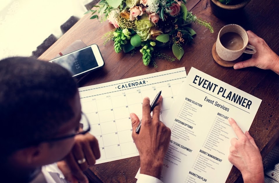 Top-down view of a calendar and an 'Event Planner' sheet being filled on a wooden table, with coffee and a smartphone nearby.