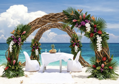 Tropical beach wedding setup with a large woven arch adorned with tropical flowers and palm leaves, over white-draped table and chairs on sand by turquoise sea.