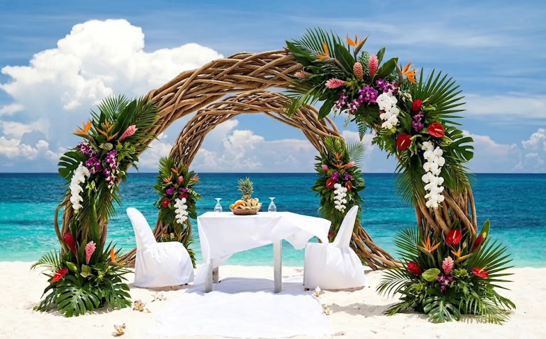 tropical beach wedding setup with a large woven arch adorned with tropical flowers and palm leaves over white draped table and chairs on sand by turquoise sea