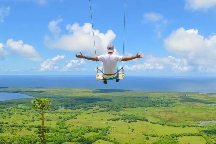 Montaña redonda miches the hidden inn punta cana the hidden inn punta cana человек на качелях, подвешенных на веревках, с высоко поднятыми руками над зеленым прибрежным пейзажем и голубым морем под ярким небом