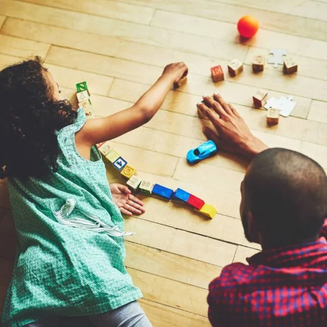 two children sit on a wooden floor arranging colorful wooden blocks into a line while a small blue toy car sits nearby