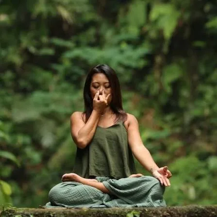 woman meditating in a forest seated crosslegged on a mossy surface with eyes closed and hands in a mudra gesture at chestface level