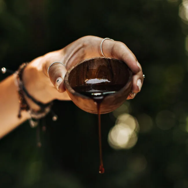 two hands cradle a small wooden cup as a dark liquid drips from its edge bracelets visible on the wrists