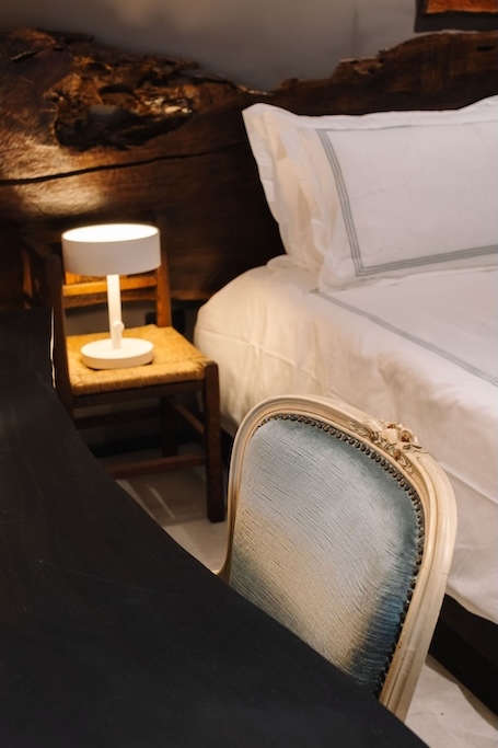 un angolo intimo della camera da letto con un letto bianco dalla testiera in legno, un piccolo comodino con una lampada e, in primo piano, una poltrona imbottita blu