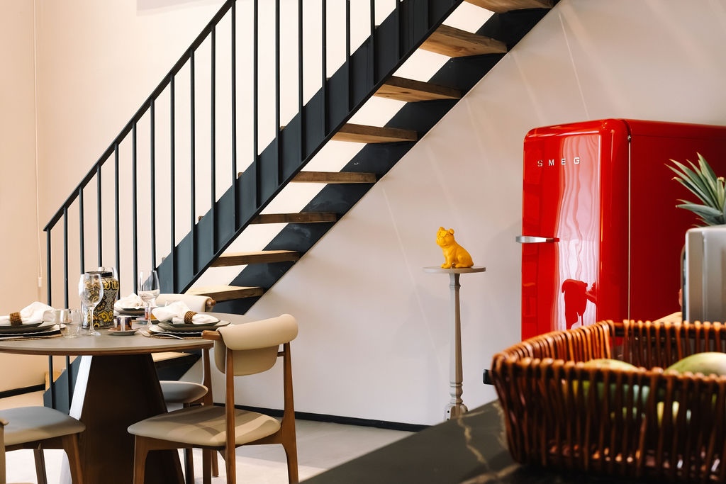 zona pranzo a pianta aperta con una scala in metallo nero, un tavolo da pranzo rotondo e un frigorifero Smeg rosso in stile retrò addossato a una parete bianca, oltre a una piccola statuetta di un cane giallo su un piedistallo vicino alle scale