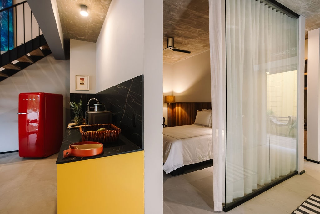 Suite d'albergo a pianta aperta con un'isola da cucina gialla, un frigorifero rosso e una zona lavello; sul bancone sono appoggiati un cesto di vimini e un ananas, mentre oltre le porte a vetri e le tende trasparenti si intravede la camera da letto