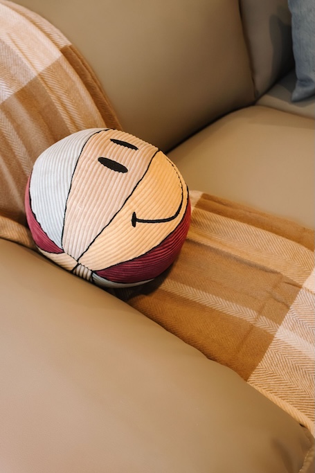 Una palla di stoffa con faccine sorridenti, con inserti color pesca chiaro e rosso, è appoggiata su un divano beige accanto a una coperta a righe arancione