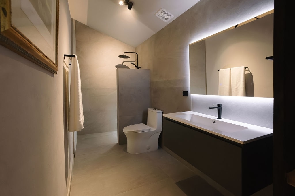 bagno moderno con lavabo bianco a parete, rubinetto nero, specchio retroilluminato, pareti beige, portasciugamani e WC accanto alla zona doccia