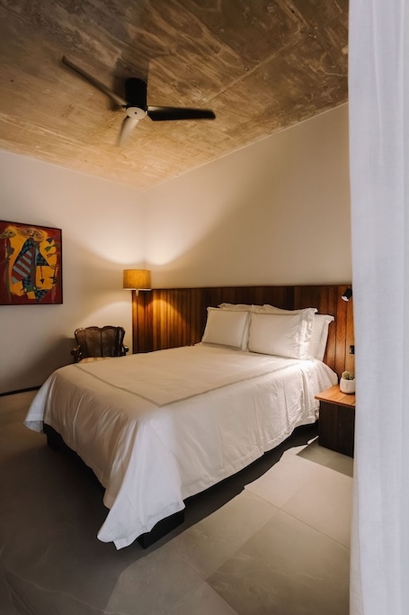 camera da letto in stile alberghiero con un letto coperto da un piumone bianco, testiera in legno e lampade su ciascun comodino; un ventilatore a soffitto pende dall’alto in uno spazio illuminato da luci calde