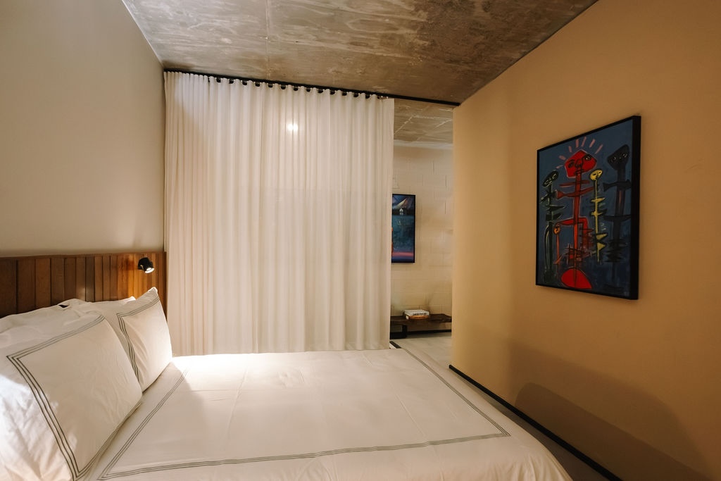 camera d'albergo con un grande letto, lenzuola bianche e una testiera in legno, accanto a una parete beige e a una piccola lampada da lettura