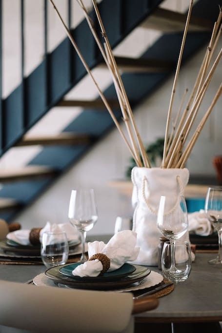 Mesa de restaurante puesta con platos, servilletas en servilleteros, copas de vino y un centro de mesa alto de caña en un jarrón blanco