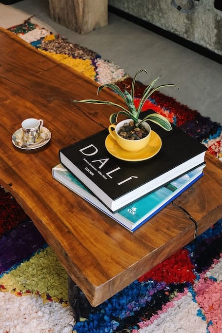 una mesa de centro de madera con una pequeña planta en maceta sobre un platillo amarillo, encima de un libro de Dalí; cerca, a la izquierda, sobre una alfombra de colores, hay una taza de té y un platillo.
