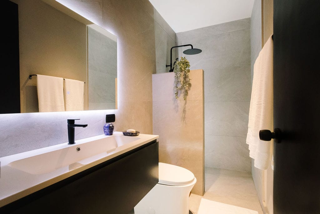 Un cuarto de baño moderno con un lavabo doble blanco, un grifo negro, un espejo retroiluminado y un inodoro situado bajo una pared alicatada en gris claro; una ducha a ras de suelo con un cabezal de ducha tipo lluvia y una planta colgante; toallas de color beige en un toallero cercano.