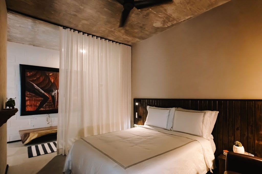 Dormitorio de estilo hotelero con una cama blanca, cabecero de madera oscura y cortinas blancas transparentes que separan los espacios.