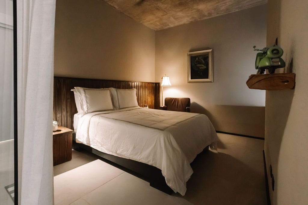 Habitación de hotel con una cama grande con sábanas blancas, cabecero de madera, una lámpara de mesilla y un scooter vintage verde en una estantería de pared.