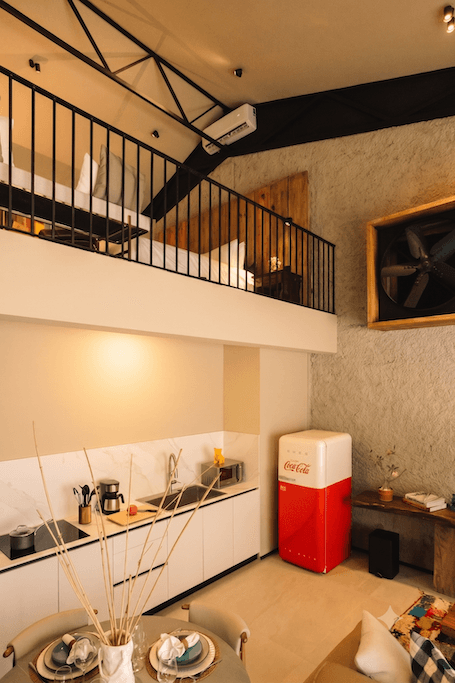 Cocina de estilo loft situada debajo de un altillo, con armarios blancos, salpicadero de mármol y una mininevera roja de Coca-Cola.