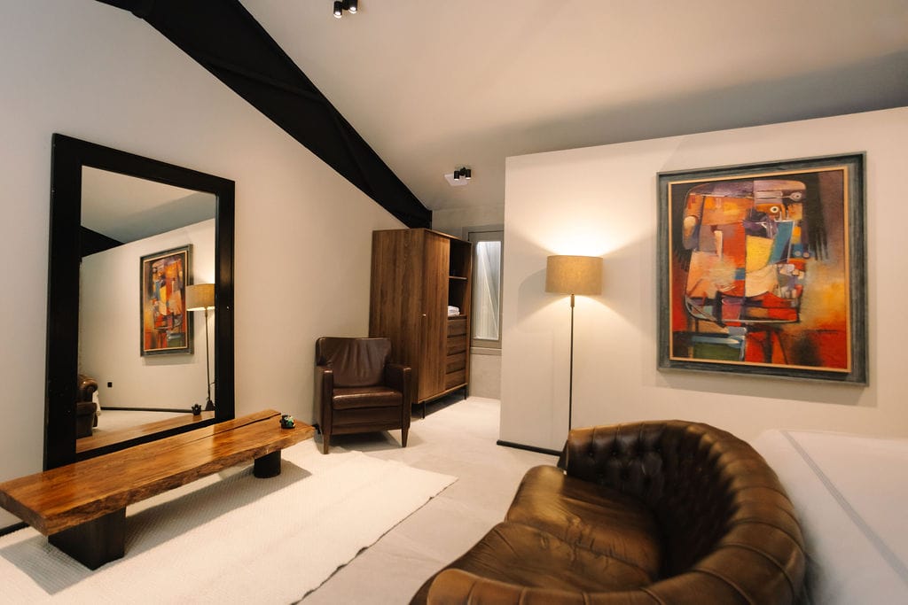 Salón contemporáneo con un sillón de cuero marrón, un sofá capitoné, un banco de madera y un gran espejo con marco negro en el que se refleja una obra de arte; salón moderno con un armario de madera, una lámpara de pie y un colorido cuadro abstracto en la pared.