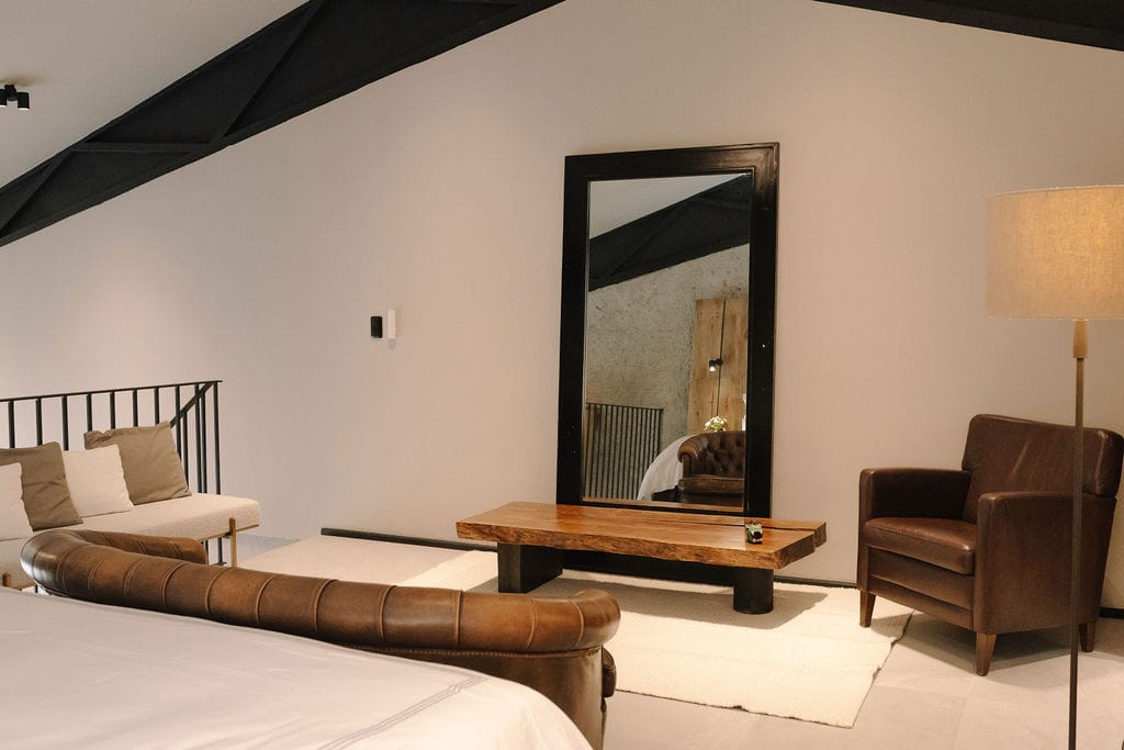 Dormitorio de estilo loft con vigas inclinadas a la vista, un gran espejo con marco negro, una mesa de centro rústica de madera, un banco de cuero marrón a los pies de la cama y un sillón de cuero marrón con una lámpara de pie junto a él, sobre una alfombra blanca.