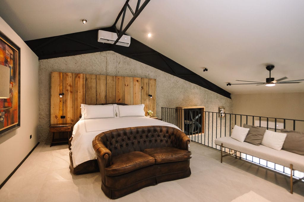Coke room main bed the hidden inn punta cana loft style bedroom with a white bed against a rustic wooden headboard a brown tufted leather sofa at the foot and a beige lounge sofa by a railing