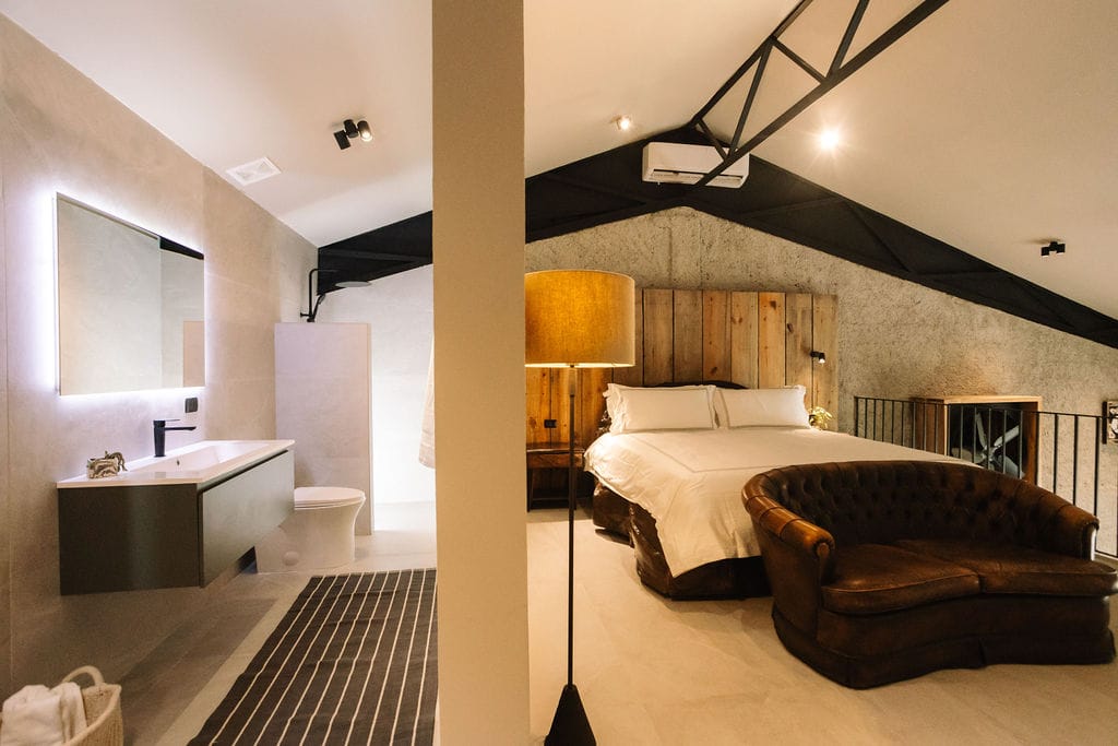 Habitación de hotel tipo loft con una cama king size, un sofá de cuero capitoné, un cabecero de madera y un cuarto de baño privado a la izquierda