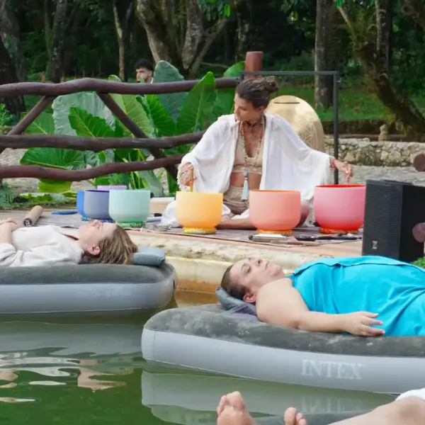outdoor meditation scene a woman in a white robe plays colorful singing bowls while two people rest on inflatable pool loungers in a pond under leafy greenery