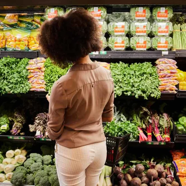 Vista de espaldas de una mujer con el pelo rizado que está eligiendo verduras de hoja verde en la sección de frutas y verduras de un supermercado; se ve una cesta de la compra a su lado.