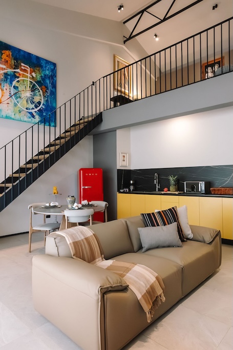 zona giorno in stile loft moderno con un divano beige, copriletti a quadri e un frigorifero rosso in stile retrò sotto la ringhiera della scala che porta al soppalco