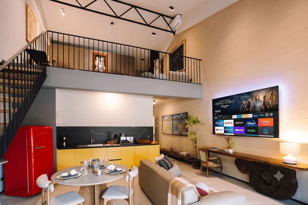 Salón diáfano con un sofá beige, una mesa de comedor redonda, armarios de cocina amarillos y un gran televisor inteligente montado en la pared en el que se pueden ver aplicaciones de streaming.