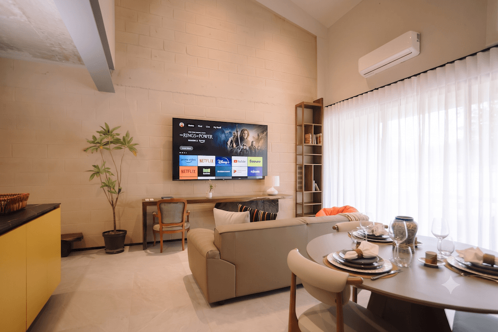 soggiorno in stile contemporaneo con una TV a parete, un divano componibile beige e un tavolo da pranzo rotondo apparecchiato per un pasto, accanto a finestre con tende trasparenti