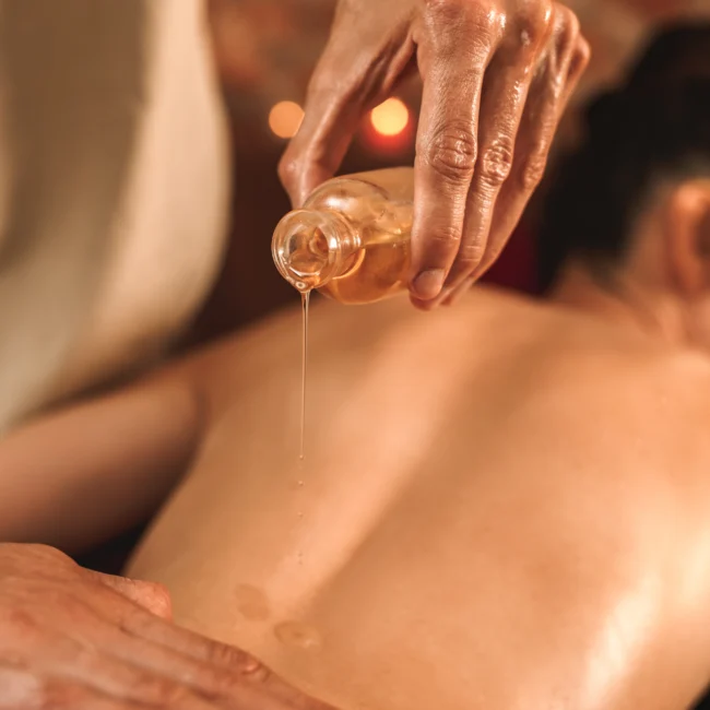 Un masajista que vierte aceite de masaje caliente de una botella pequeña sobre la espalda desnuda de una persona durante un tratamiento de spa