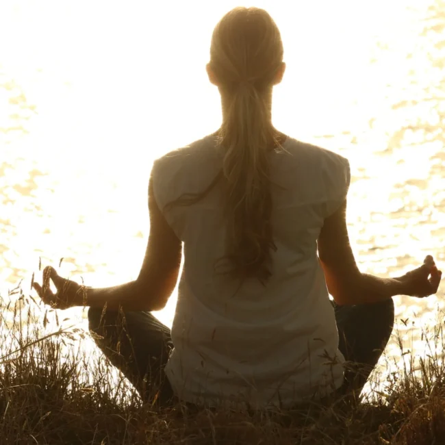 Una persona con el pelo largo recogido en una coleta está sentada con las piernas cruzadas a orillas de un lago, meditando al atardecer.