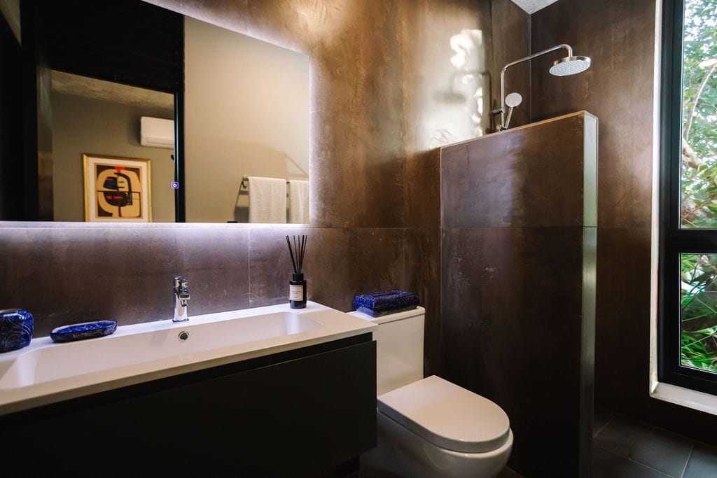 Private loft bathroom the hidden inn punta cana modern bathroom with a long white counter sink dark cabinetry brown metallic walls a glass shower to the right and a large window showing greenery