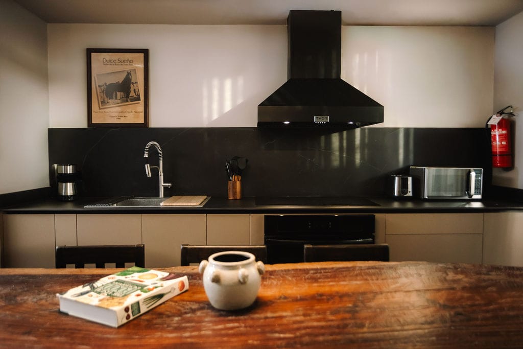 Private loft kitchen the hidden inn punta cana modern kitchen with black countertops and backsplash stainless sink stove with large range hood microwave and toaster along the counter and a wooden table in the foreground with a book and a ceramic pot