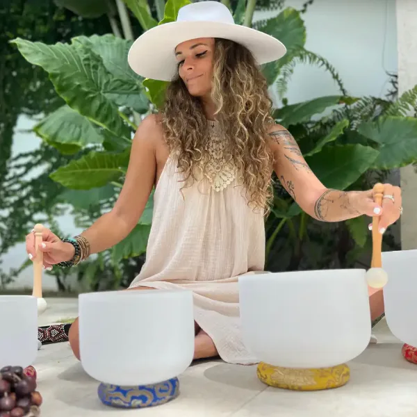 woman in a wide brimmed white hat sits cross legged outdoors playing crystal singing bowls with wooden mallets in a tropical garden