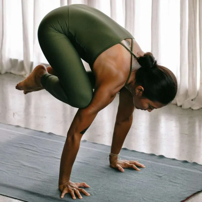 woman in olive green leggings in crow pose on a blue yoga mat indoors