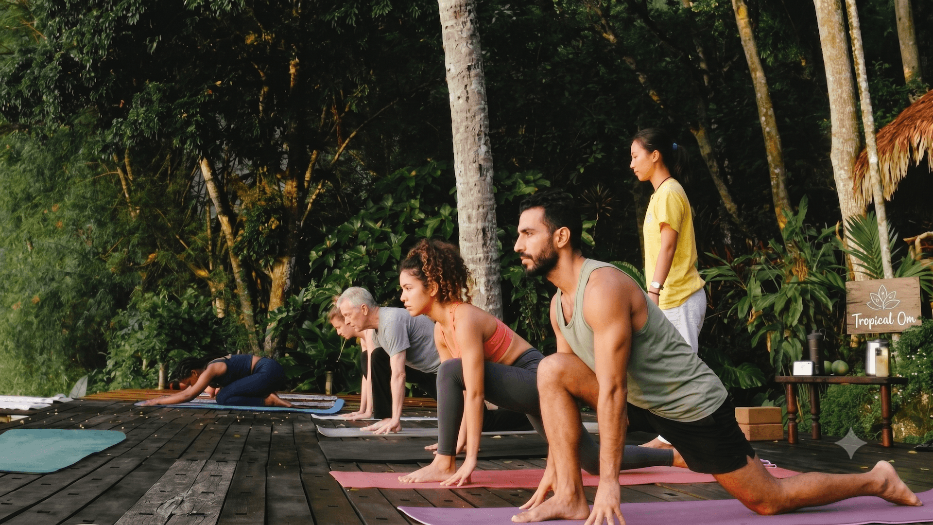 Personas practicando yoga sobre colchonetas en una terraza de madera, en un entorno tropical al aire libre.